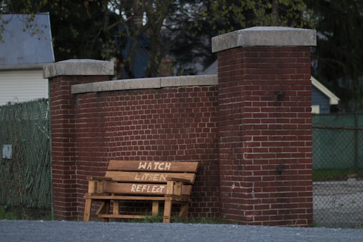 A wooden bench with the words 'WATCH LISTEN REFLECT' painted on it is placed against a tall red brick wall between two pillars. The setting includes a chain link fence with some foliage in the background, and a house partially visible in the distance.
