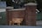 A wooden bench with the words 'WATCH LISTEN REFLECT' painted on it is placed against a tall red brick wall between two pillars. The setting includes a chain link fence with some foliage in the background, and a house partially visible in the distance.