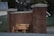 A wooden bench with the words 'WATCH LISTEN REFLECT' painted on it is placed against a tall red brick wall between two pillars. The setting includes a chain link fence with some foliage in the background, and a house partially visible in the distance.