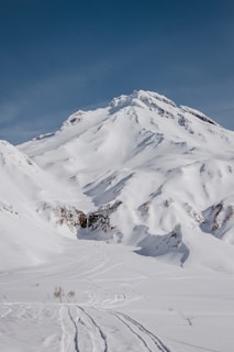 A snow-covered mountain peak rises against a clear blue sky, with undisturbed snow slopes and rocky outcrops visible. Tracks in the snow suggest ski or snowboard activities.