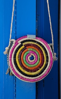 A round, woven bag with a colorful spiral pattern hangs against a vibrant blue background. The bag features concentric circles in shades of pink, orange, yellow, green, and blue, with a rope strap attached.