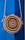 A round, woven bag with a colorful spiral pattern hangs against a vibrant blue background. The bag features concentric circles in shades of pink, orange, yellow, green, and blue, with a rope strap attached.