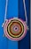 A round, woven bag with a colorful spiral pattern hangs against a vibrant blue background. The bag features concentric circles in shades of pink, orange, yellow, green, and blue, with a rope strap attached.