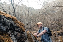 A man wearing a cap, t-shirt, and backpack explores a moss-covered rocky terrain in a sparsely vegetated, dry forest area. He is focused on the rocks in front of him, holding a camera with a strap over his shoulder.