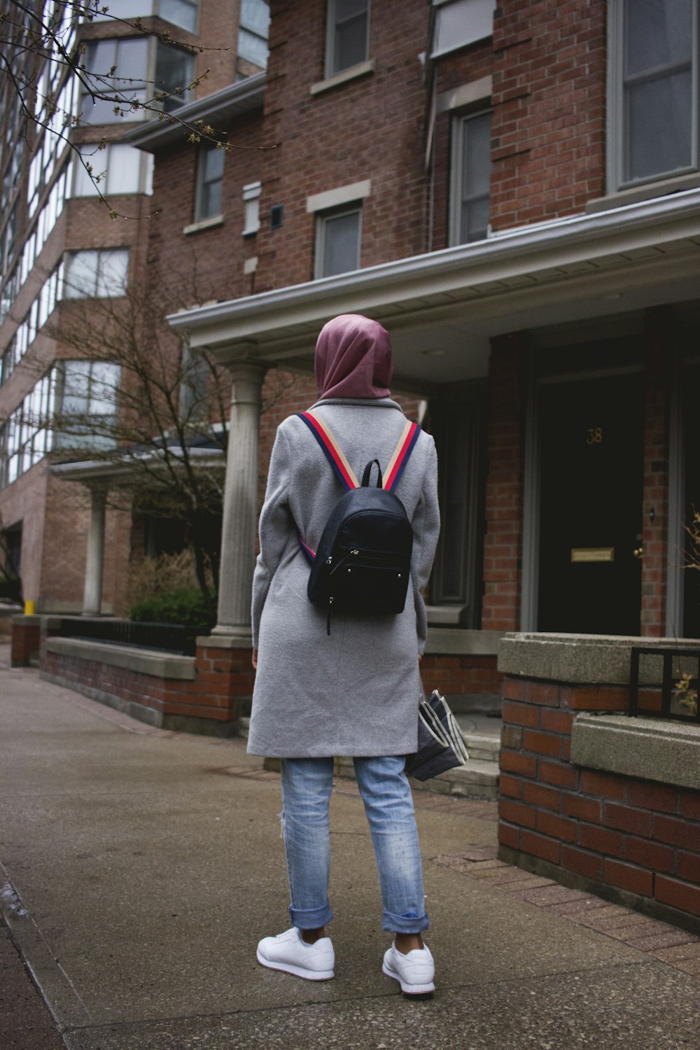 woman in pink hijab headdress, gray coat, blue jeans, white shoes, and black backpack standing near house
