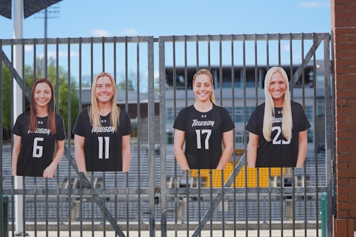 Four cutout images of people are mounted on a metal fence with a sports stadium in the background.