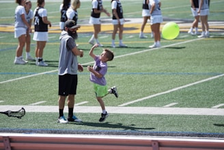 A young child and an adult are playing on a sports field. The child is wearing a purple shirt and lime green shorts, reaching up towards a bright green balloon. The adult, in athletic wear, is standing nearby, appearing engaged. In the background, more people in uniforms are standing, possibly indicating a team setting.