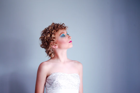 A person with curly hair styled elegantly, wearing a strapless white dress with sequins. The makeup is bold, featuring bright blue eye shadow and pink lipstick. The person gazes upward with a serene expression against a plain, soft blue background.