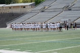 A line of female athletes stands on a sports field, each wearing white uniforms with numbers on their backs. They appear to be engaged in a pre-game or ceremonial activity. The background shows empty stadium seating, indicating it's an outdoor venue with a sports track.