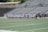 A line of female athletes stands on a sports field, each wearing white uniforms with numbers on their backs. They appear to be engaged in a pre-game or ceremonial activity. The background shows empty stadium seating, indicating it's an outdoor venue with a sports track.