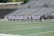 A line of female athletes stands on a sports field, each wearing white uniforms with numbers on their backs. They appear to be engaged in a pre-game or ceremonial activity. The background shows empty stadium seating, indicating it's an outdoor venue with a sports track.