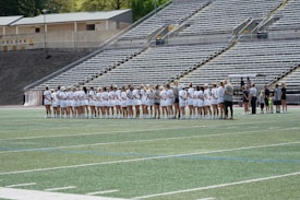A line of female athletes stands on a sports field, each wearing white uniforms with numbers on their backs. They appear to be engaged in a pre-game or ceremonial activity. The background shows empty stadium seating, indicating it's an outdoor venue with a sports track.