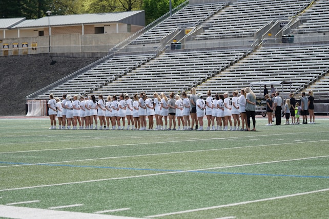 A line of female athletes stands on a sports field, each wearing white uniforms with numbers on their backs. They appear to be engaged in a pre-game or ceremonial activity. The background shows empty stadium seating, indicating it's an outdoor venue with a sports track.