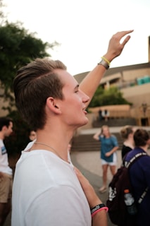 A young person with short hair is raising their hand enthusiastically in an outdoor setting. They are wearing a white shirt and several wristbands. In the background, blurred figures of other people are visible, likely suggesting a group gathering or event.