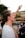 A young person with short hair is raising their hand enthusiastically in an outdoor setting. They are wearing a white shirt and several wristbands. In the background, blurred figures of other people are visible, likely suggesting a group gathering or event.