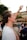 A young person with short hair is raising their hand enthusiastically in an outdoor setting. They are wearing a white shirt and several wristbands. In the background, blurred figures of other people are visible, likely suggesting a group gathering or event.