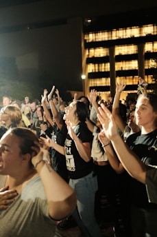 A group of people raising their hands, appearing to be in a moment of unity or celebration. The event seems to be taking place outdoors at night, with an illuminated building in the background providing a warm glow. Many individuals are wearing casual clothing, and a few have expressions of joy or seriousness.