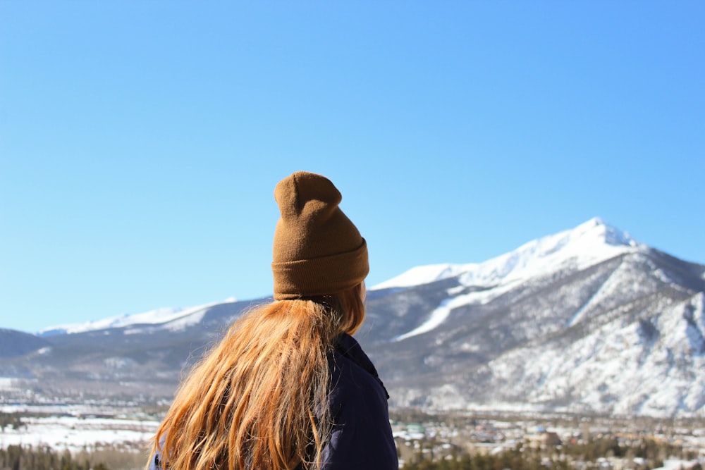 person with blonde hair and brown beanie hat facing mountain