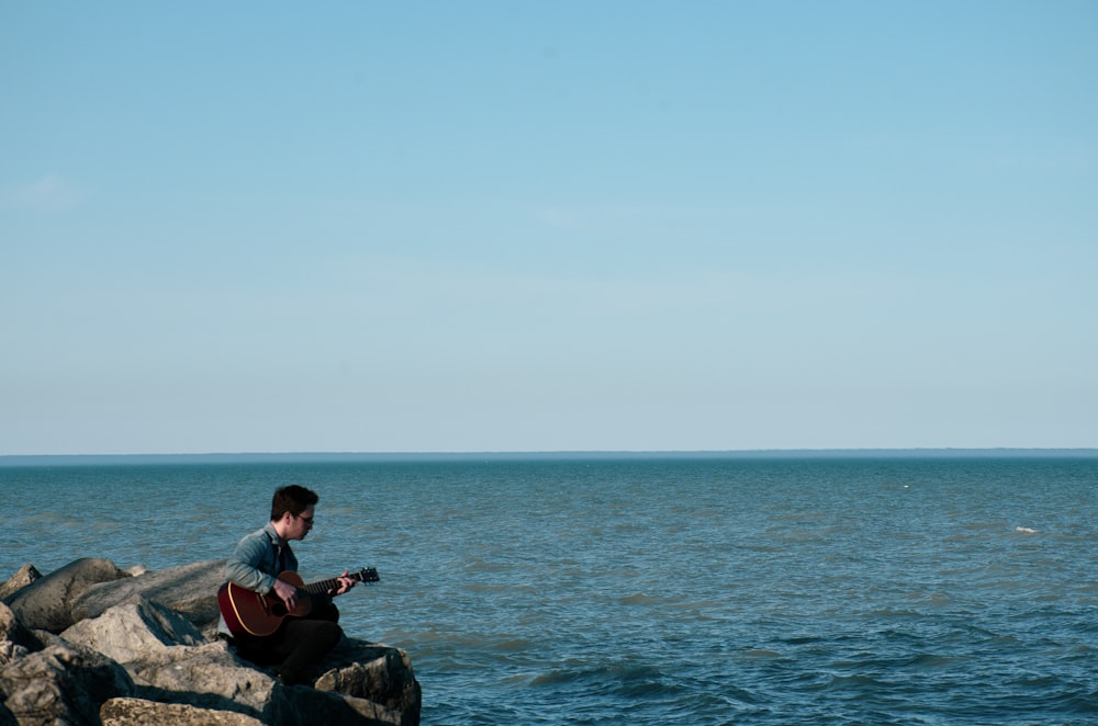 man playing guitar sitting on rock near ocean