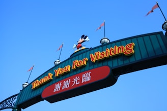 An archway sign with the phrase 'Thank You For Visiting' in prominent yellow letters. Below, there is text in Chinese. A character wearing red shorts and yellow shoes stands on top of the arch, and the sky is clear blue in the background.