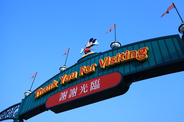 An archway sign with the phrase 'Thank You For Visiting' in prominent yellow letters. Below, there is text in Chinese. A character wearing red shorts and yellow shoes stands on top of the arch, and the sky is clear blue in the background.