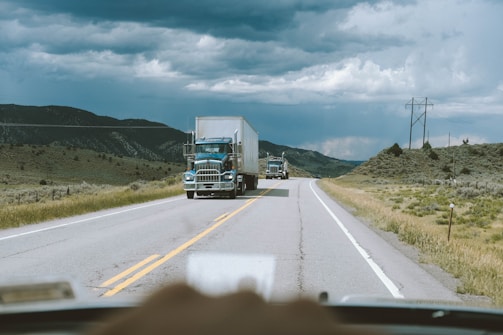 A highway scene with large trucks traveling on a two-lane road. The landscape is rural and mountainous, with grassy hills and distant mountains under a cloudy sky. The perspective is from inside a vehicle, with a hint of a dashboard visible at the bottom.