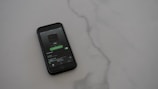 A smartphone with a dark case displays a music streaming app on the screen, placed on a light gray, smooth surface. The app interface is showing an album cover and music controls.