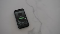 A smartphone with a dark case displays a music streaming app on the screen, placed on a light gray, smooth surface. The app interface is showing an album cover and music controls.
