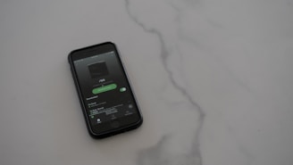 A smartphone with a dark case displays a music streaming app on the screen, placed on a light gray, smooth surface. The app interface is showing an album cover and music controls.