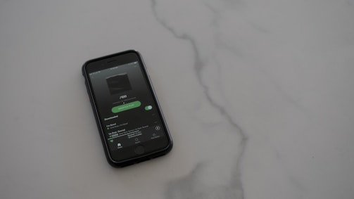 A smartphone with a dark case displays a music streaming app on the screen, placed on a light gray, smooth surface. The app interface is showing an album cover and music controls.