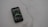 A smartphone with a dark case displays a music streaming app on the screen, placed on a light gray, smooth surface. The app interface is showing an album cover and music controls.