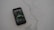 A smartphone with a dark case displays a music streaming app on the screen, placed on a light gray, smooth surface. The app interface is showing an album cover and music controls.