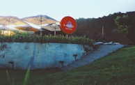 A bright outdoor setting features several large white umbrellas providing shade over a modern seating area. A prominent circular red sign with fish imagery and text stands out against the greenery. The scene is bordered by a stone wall with lush plants growing on top and a staircase leading up the hill covered in green grass.