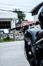 A close-up view of a sleek black motorcycle, focusing on the handlebars and rearview mirror. The background includes a blurred perspective of a road and buildings with greenery. The image captures the motorcycle's glossy surface and detailed mechanics.