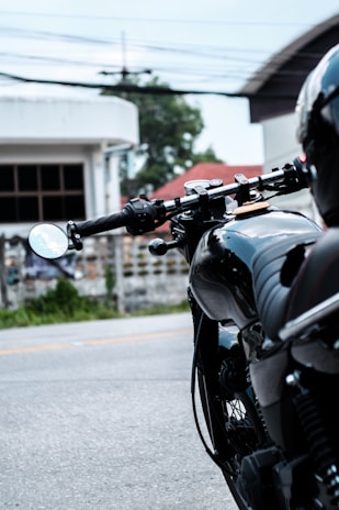 A close-up view of a sleek black motorcycle, focusing on the handlebars and rearview mirror. The background includes a blurred perspective of a road and buildings with greenery. The image captures the motorcycle's glossy surface and detailed mechanics.
