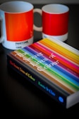 A book titled 'The Secret Lives of Colour' with a vibrant cover featuring various colorful stripes is placed on a dark surface. Next to it are two ceramic mugs with a color gradient design, labeled with Pantone color codes.