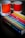 A book titled 'The Secret Lives of Colour' with a vibrant cover featuring various colorful stripes is placed on a dark surface. Next to it are two ceramic mugs with a color gradient design, labeled with Pantone color codes.