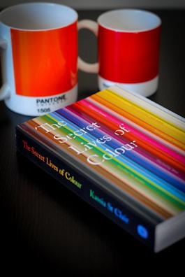 A book titled 'The Secret Lives of Colour' with a vibrant cover featuring various colorful stripes is placed on a dark surface. Next to it are two ceramic mugs with a color gradient design, labeled with Pantone color codes.