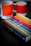 A book titled 'The Secret Lives of Colour' with a vibrant cover featuring various colorful stripes is placed on a dark surface. Next to it are two ceramic mugs with a color gradient design, labeled with Pantone color codes.