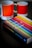 A book titled 'The Secret Lives of Colour' with a vibrant cover featuring various colorful stripes is placed on a dark surface. Next to it are two ceramic mugs with a color gradient design, labeled with Pantone color codes.
