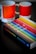 A book titled 'The Secret Lives of Colour' with a vibrant cover featuring various colorful stripes is placed on a dark surface. Next to it are two ceramic mugs with a color gradient design, labeled with Pantone color codes.