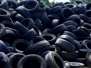 A large pile of old, used tires stacked haphazardly outdoors, with some vegetation visible in the background.