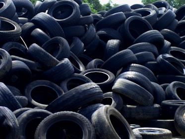 A large pile of old, used tires stacked haphazardly outdoors, with some vegetation visible in the background.