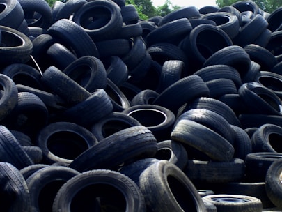 A large pile of old, used tires stacked haphazardly outdoors, with some vegetation visible in the background.