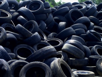 A large pile of old, used tires stacked haphazardly outdoors, with some vegetation visible in the background.