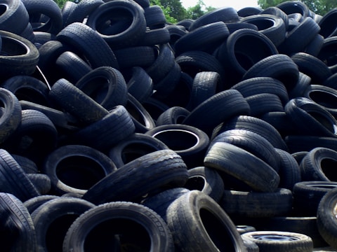 A large pile of old, used tires stacked haphazardly outdoors, with some vegetation visible in the background.