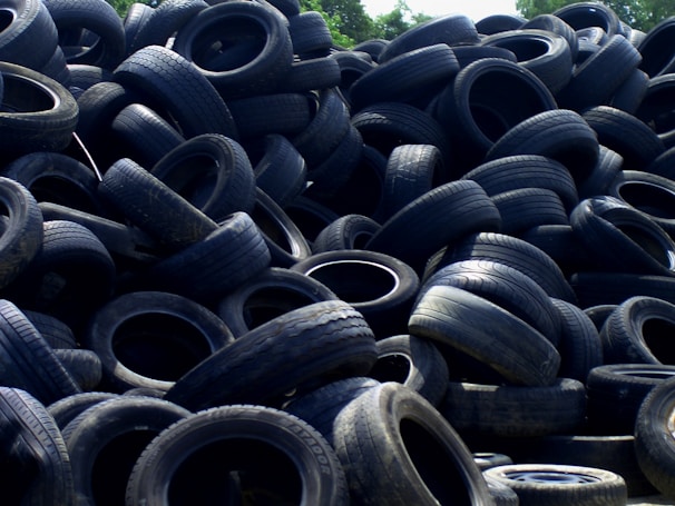 A large pile of old, used tires stacked haphazardly outdoors, with some vegetation visible in the background.