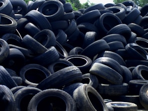 A large pile of old, used tires stacked haphazardly outdoors, with some vegetation visible in the background.