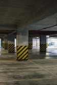 A concrete parking garage interior featuring multiple support columns with black and yellow diagonal stripes at their bases. The floor is clean and smooth, and fluorescent lights provide illumination. An emergency exit sign can be seen in the background.