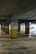 A concrete parking garage interior featuring multiple support columns with black and yellow diagonal stripes at their bases. The floor is clean and smooth, and fluorescent lights provide illumination. An emergency exit sign can be seen in the background.